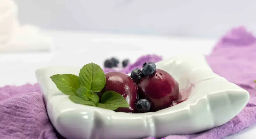 Due palline di gelato ai mirtilli, guarnite con mirtilli freschi e foglie di menta, servite in una ciotola bianca su un tovagliolo viola.