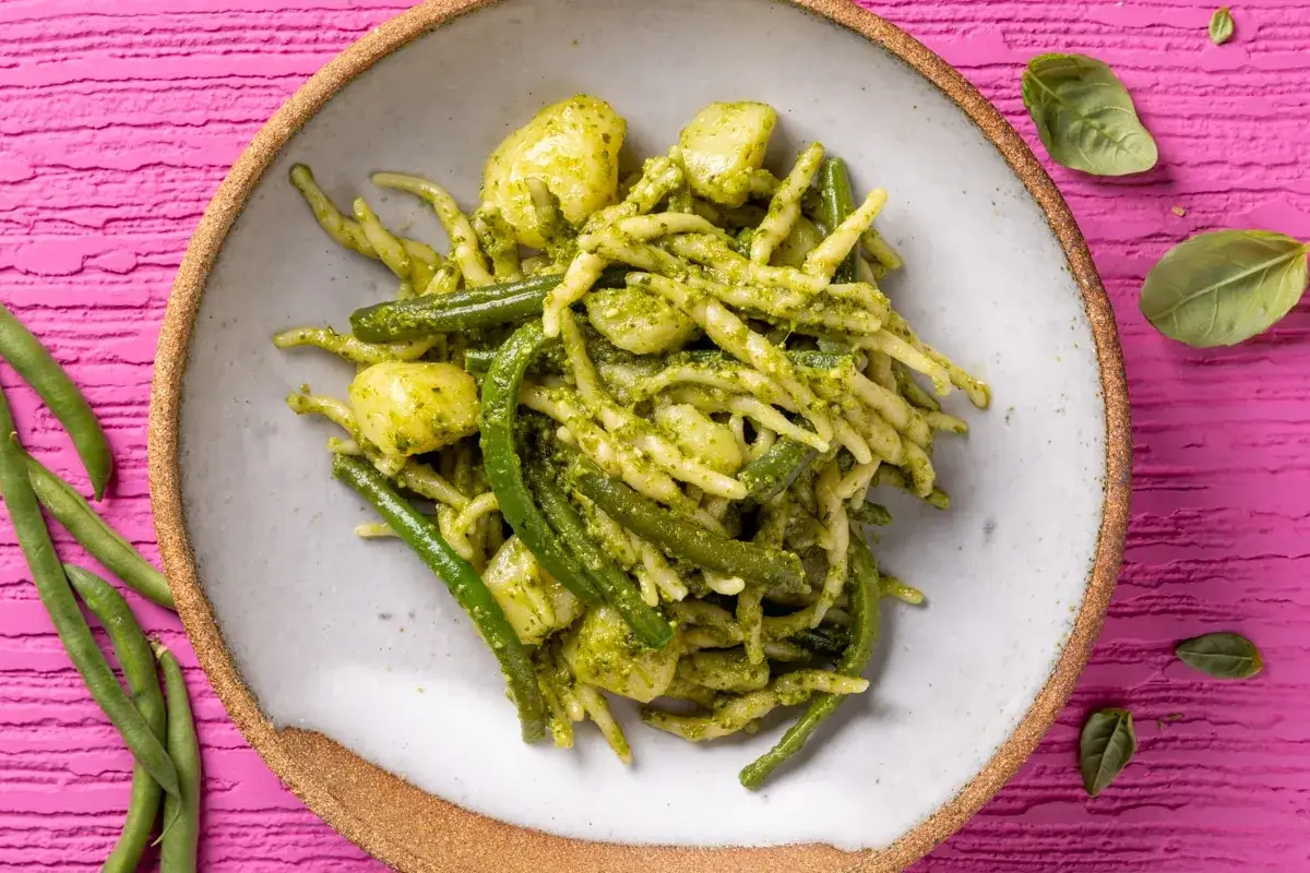 Un piatto di trofie al pesto con fagiolini e patate, su uno sfondo rosa vibrante.