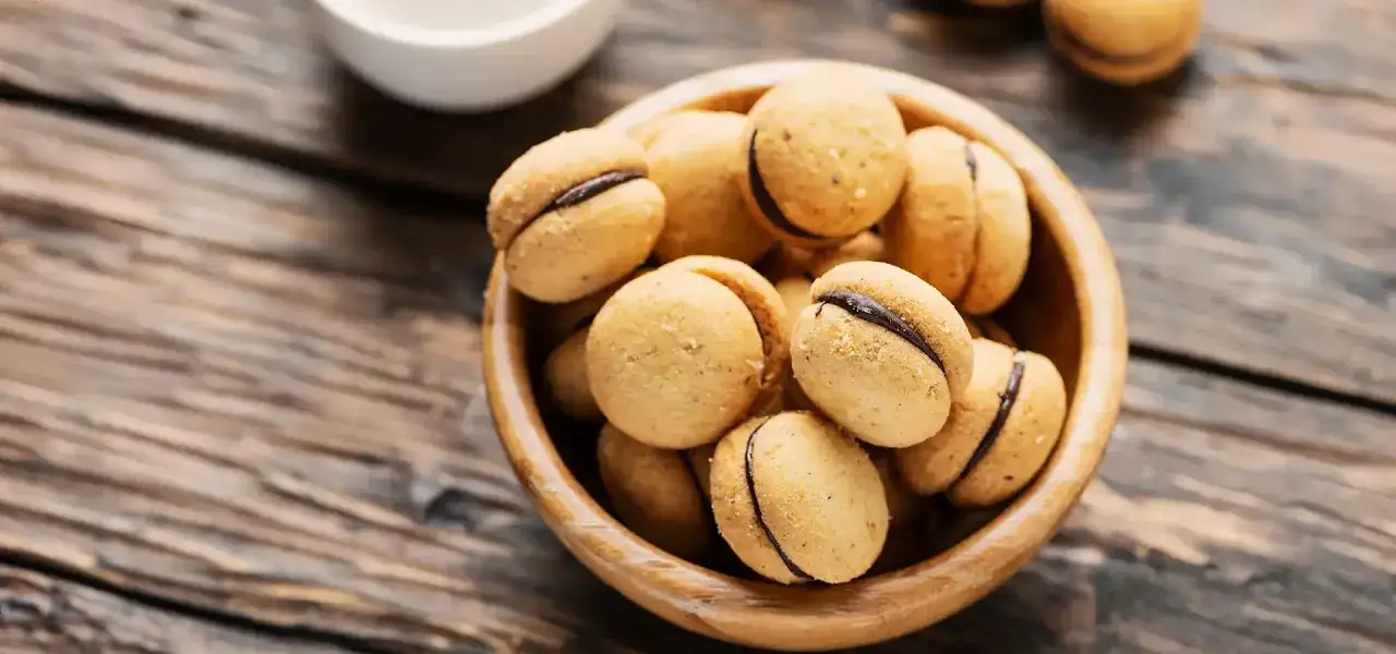 Baci di dama ricetta: biscotti friabili con ripieno di cioccolato in una ciotola di legno su un tavolo rustico.