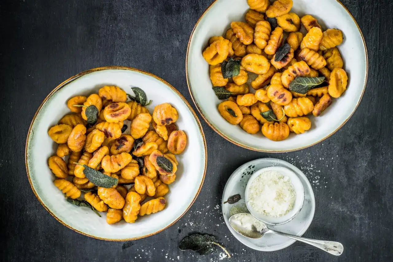 Due ciotole piene di deliziosi gnocchi di zucca, guarniti con foglie di salvia croccanti. Accanto, una ciotolina di parmigiano grattugiato.