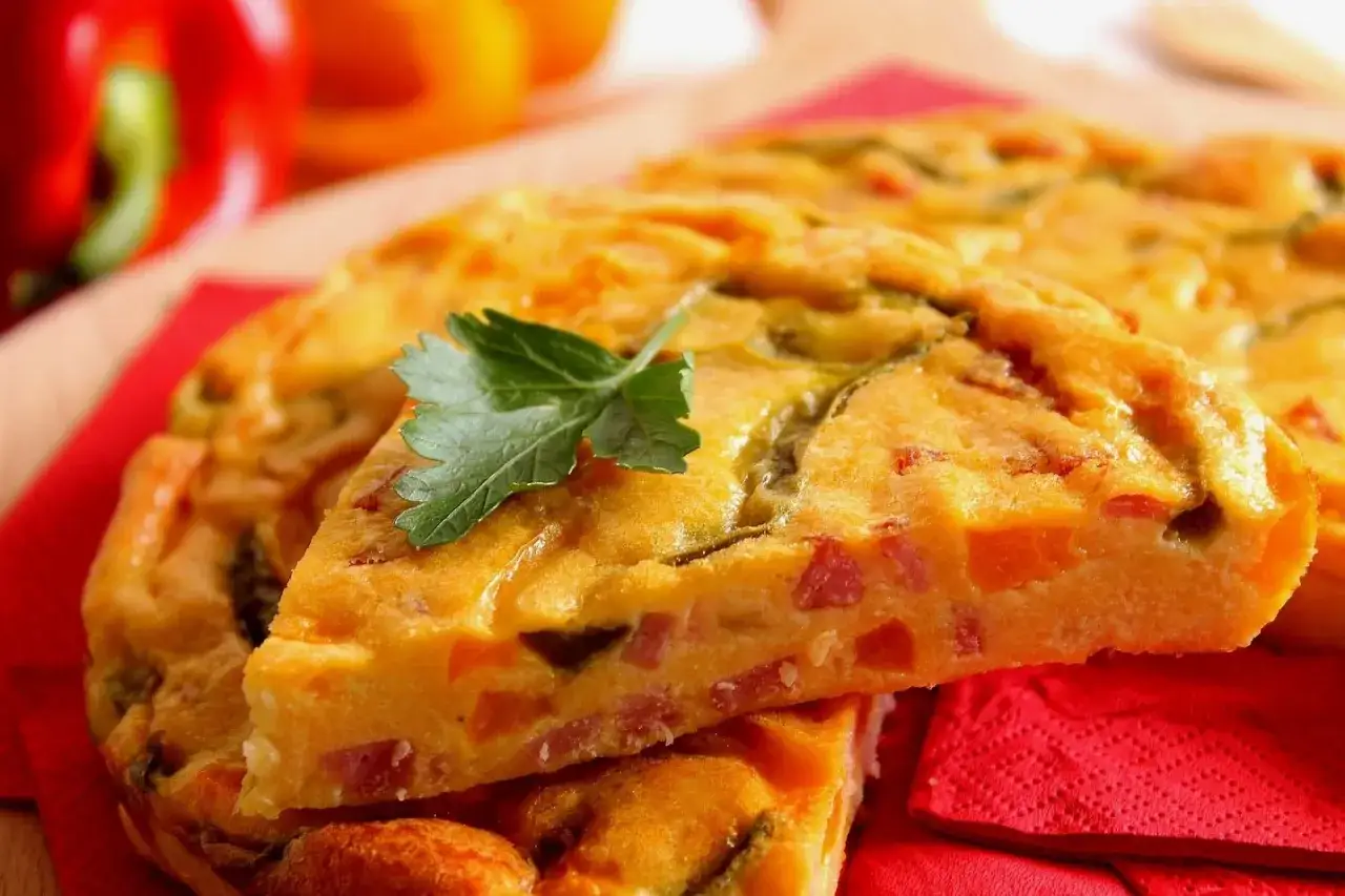 Una fetta di frittata di peperoni, ricca di verdure colorate e guarnita con prezzemolo, su un tovagliolo rosso.