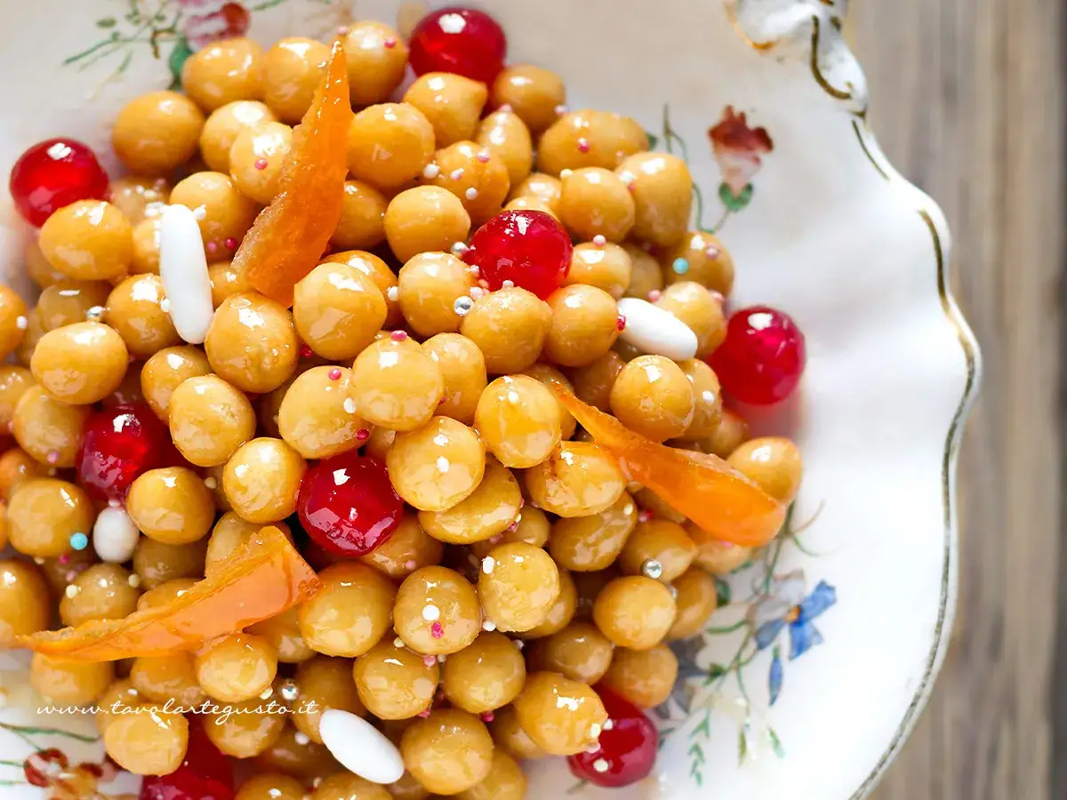 Struffoli ricetta della nonna: palline dorate e lucide, decorate con canditi rossi, bianchi e scorzette d'arancia, in un piatto floreale.