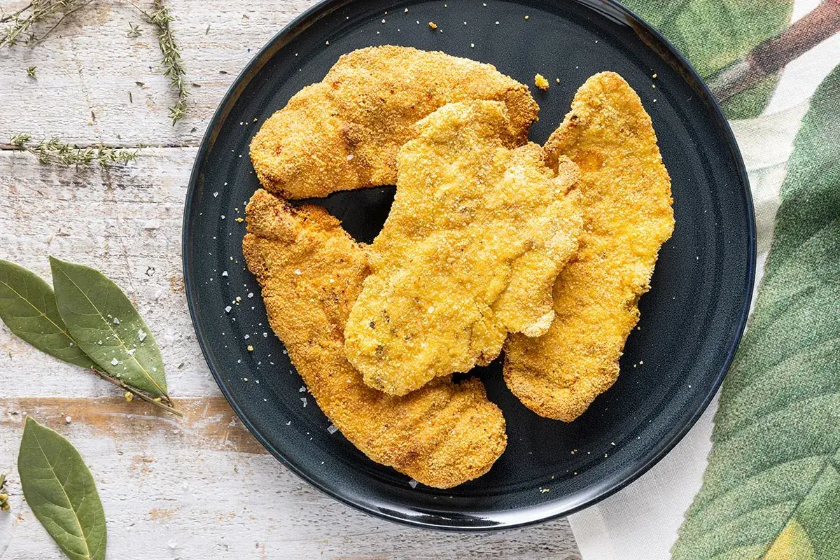 Cotoletta in friggitrice ad aria, dorata e croccante, servita su un piatto scuro con erbe aromatiche e foglie di alloro.
