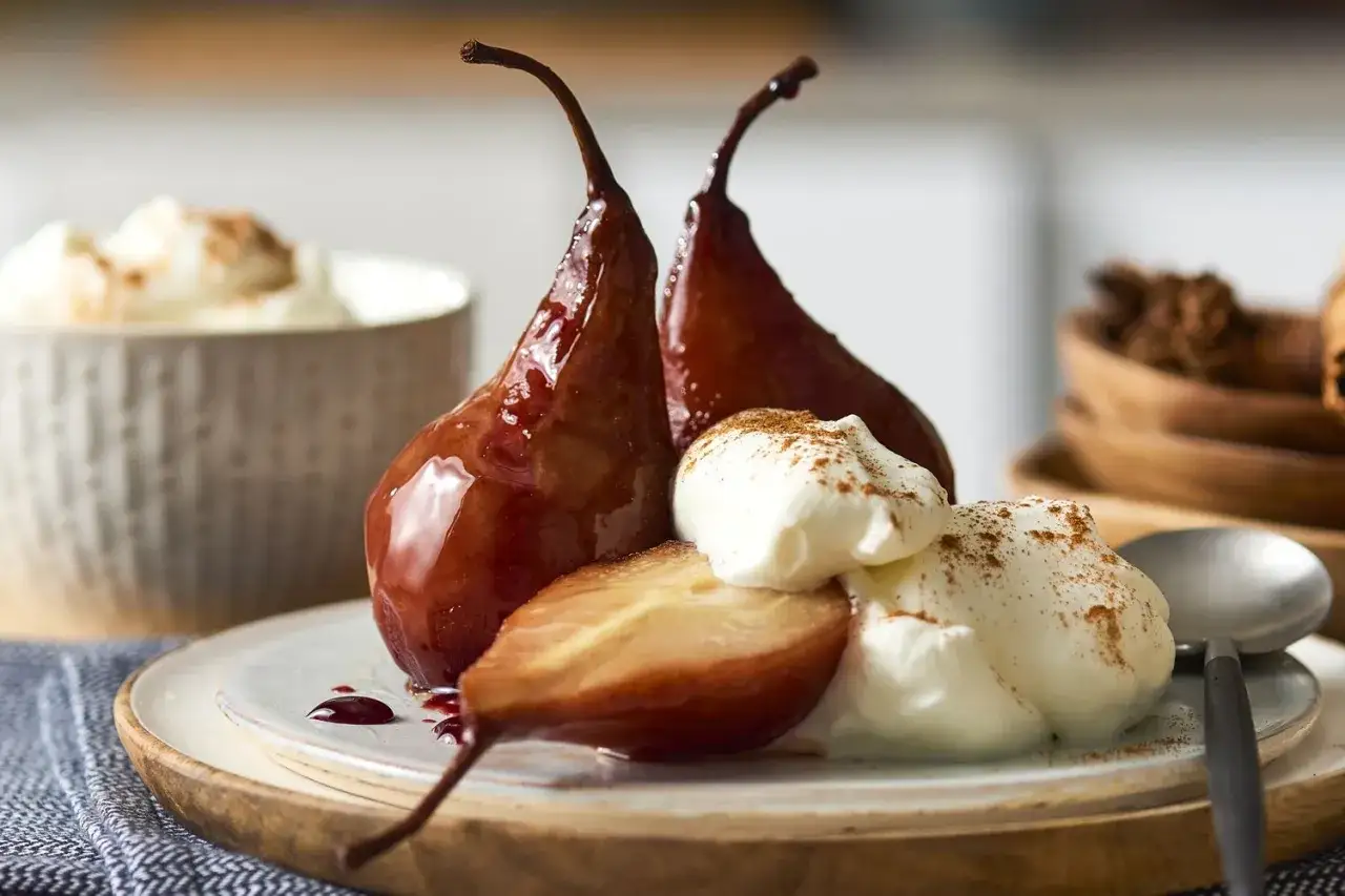 Pere cotte nel vino, lucide e scure, servite con panna montata e una spolverata di cannella. Un dolce autunnale invitante.