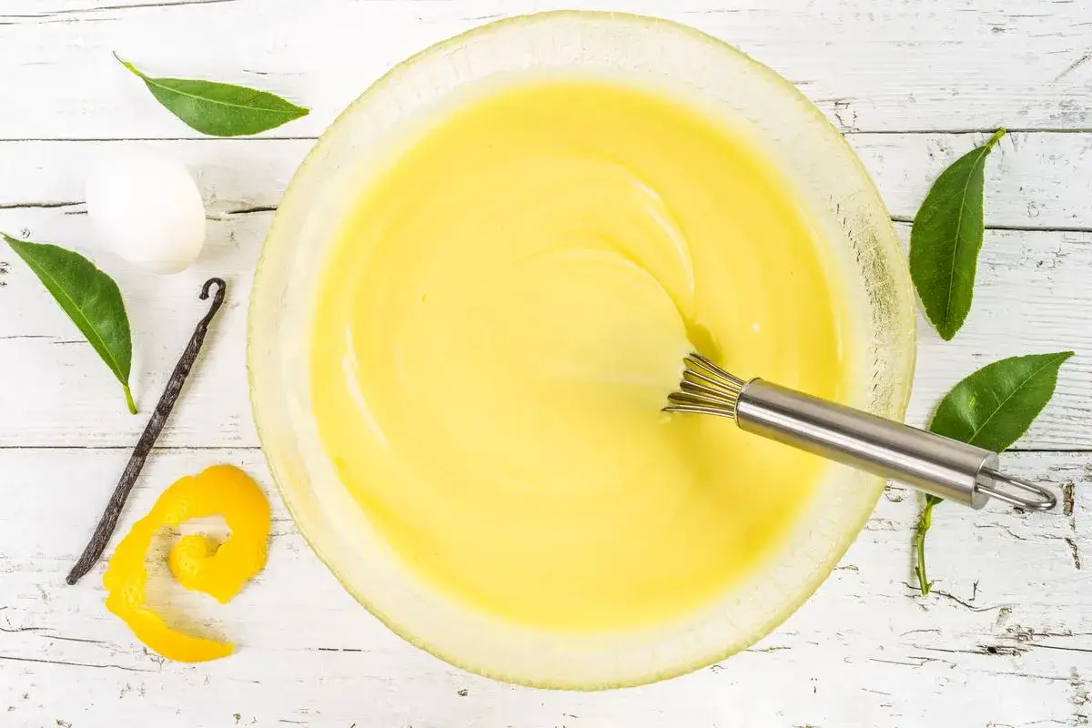 Una ciotola di uovo sbattuto con zucchero, con accanto un uovo intero, una bacca di vaniglia, scorza di limone e foglie verdi.
