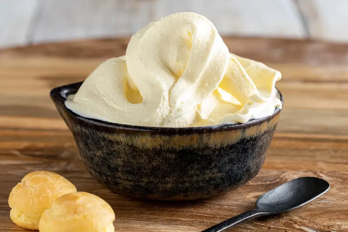 Una ciotola scura piena di cremoso gelato alla vaniglia, con due bign&egrave; e un cucchiaio scuro su un tavolo di legno.