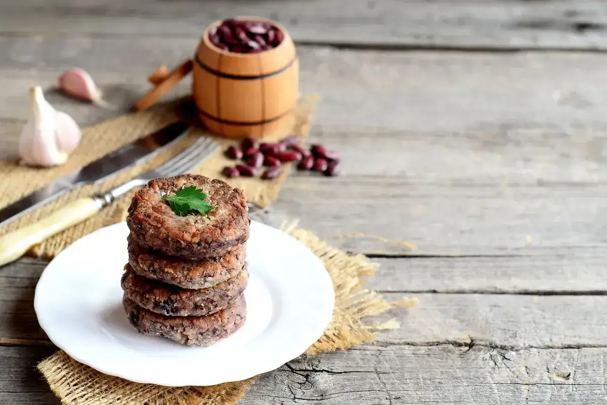 Stack di hamburger di fagioli su un piatto bianco, con fagioli rossi in un barile di legno e aglio sullo sfondo.