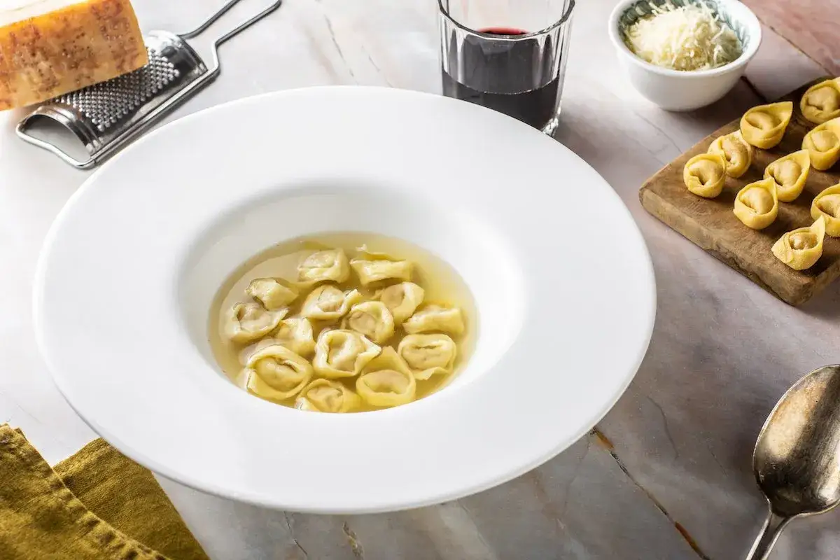 Un piatto fumante di tortellini in brodo, accompagnato da parmigiano grattugiato, un bicchiere di vino rosso e tortellini freschi su un tagliere.