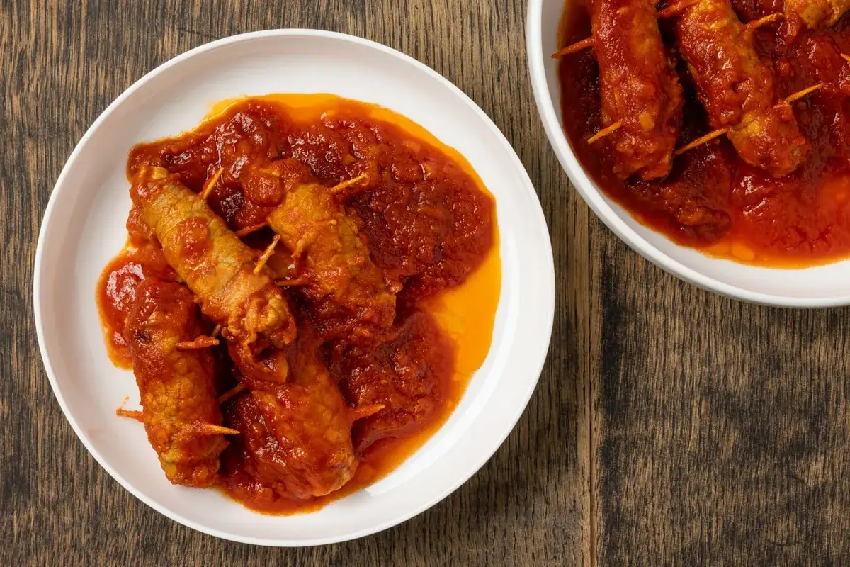Involtini alla romana succulenti in salsa rossa, serviti su piatti bianchi su un tavolo di legno rustico.