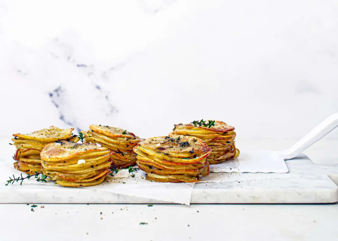 Delizioso millefoglie di patate dorate, impilate con erbe aromatiche fresche, su un tagliere di marmo.