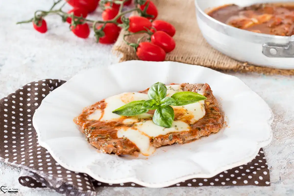 Bistecche alla pizzaiola su un piatto bianco, con mozzarella filante, pomodoro e basilico fresco. Sullo sfondo, pomodorini e una padella.