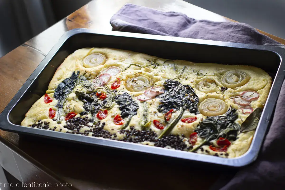 Focaccia di lenticchie appena sfornata, guarnita con cipolle, pomodorini, verdure scure e rosmarino.