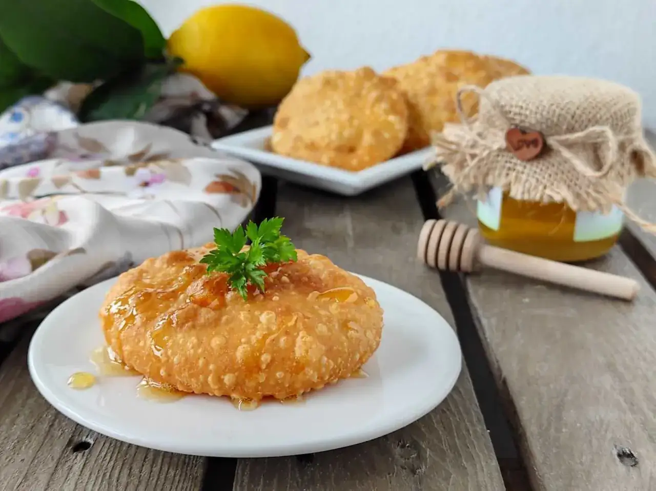 Un delizioso dolce sardo fritto, dorato e cosparso di miele, guarnito con prezzemolo fresco. Sullo sfondo, limone, altri dolci e un vasetto di miele.