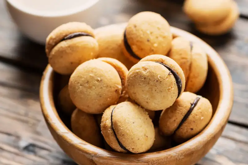 Baci di dama ricetta: deliziosi biscotti alle nocciole con ripieno di cioccolato, serviti in una ciotola di legno.