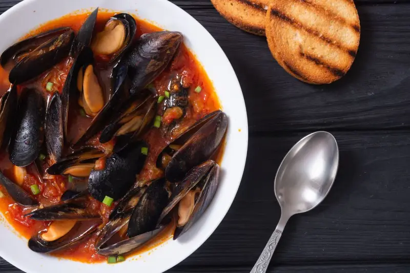 Un invitante piatto di zuppa di cozze in salsa rossa, accompagnato da crostini tostati e un cucchiaio d'argento.